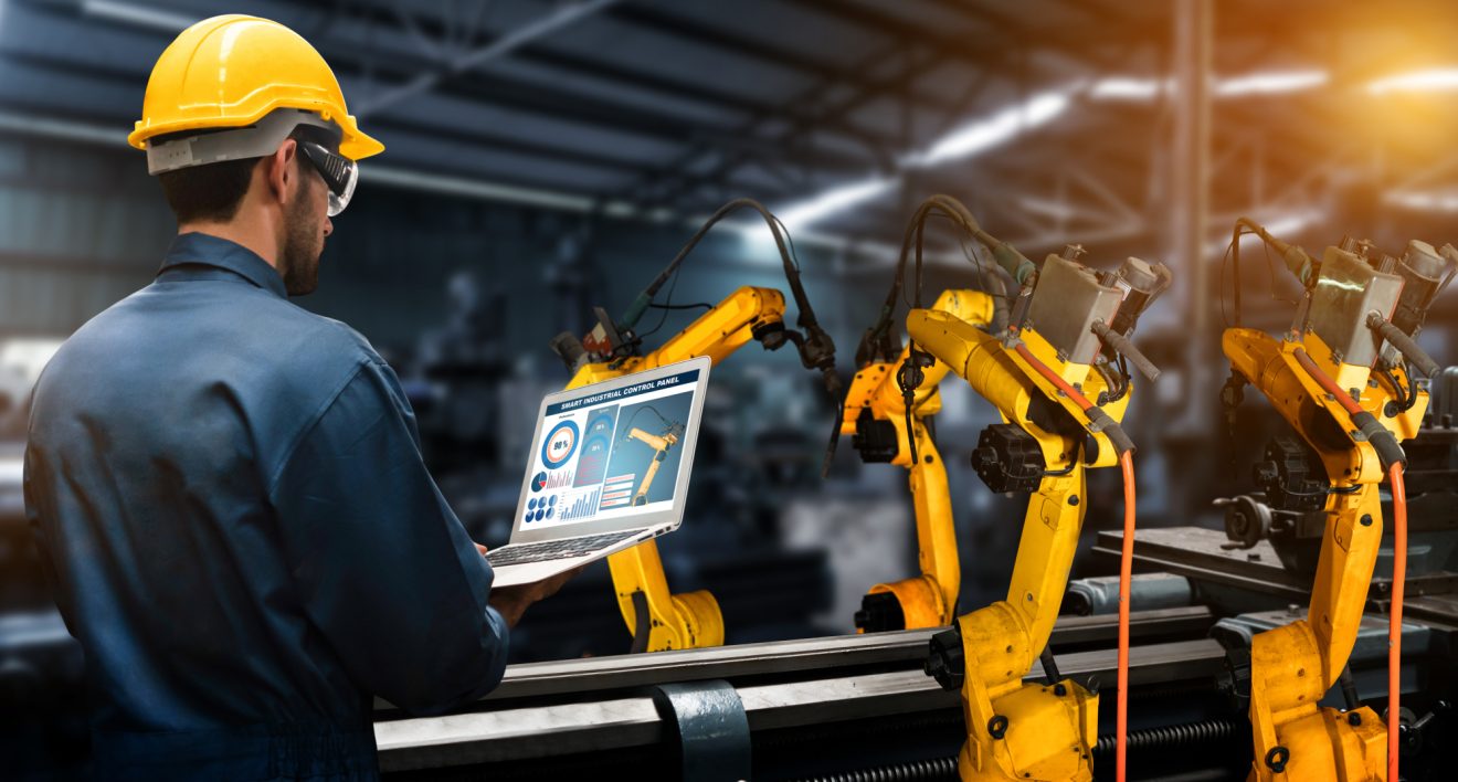 Man in yellow hardhat holding laptop while inspecting manufacturing equipment