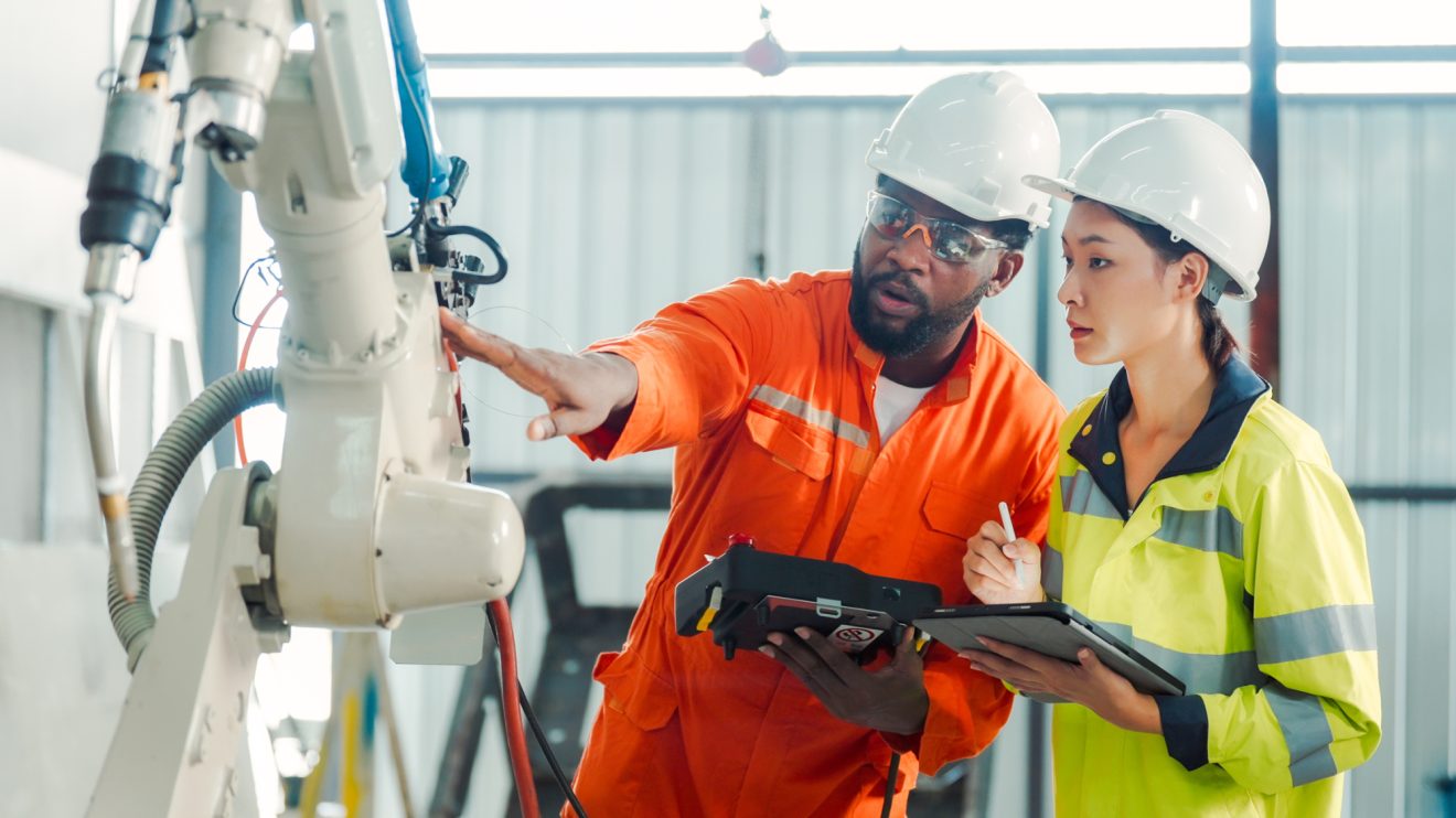 Two people in bright jumpsuits and hardhats inspect a piece of manufacturing equipment