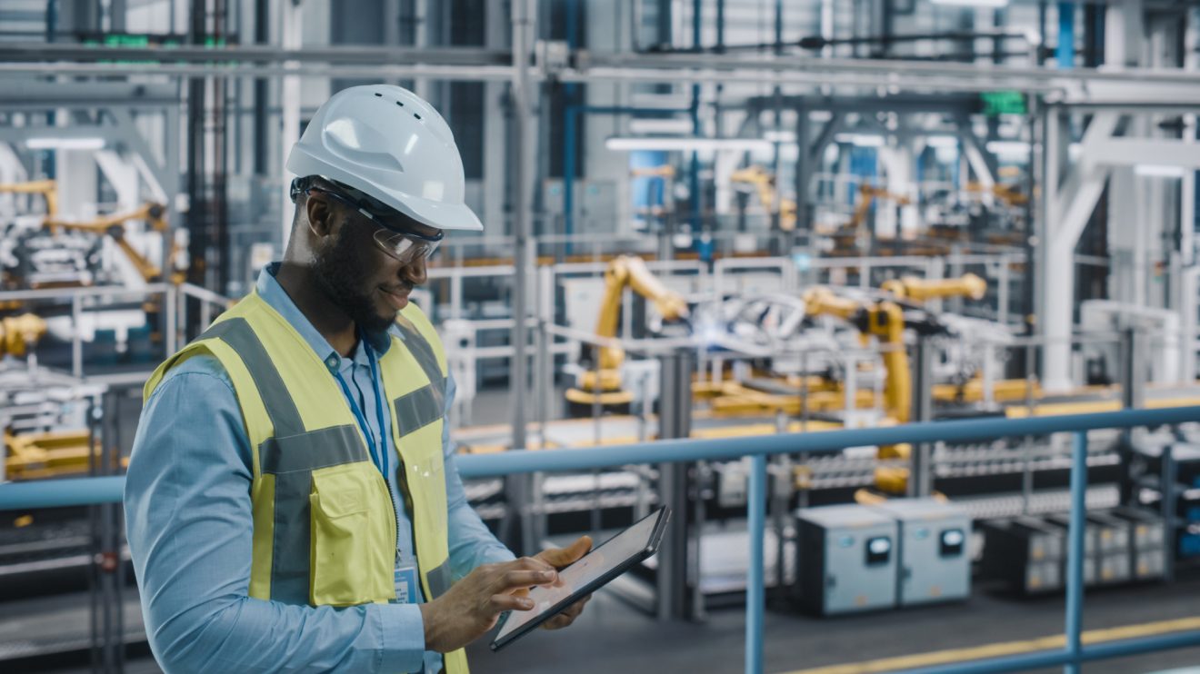 A man in a hardhat and safety vest uses a tablet in a manufacturing plant