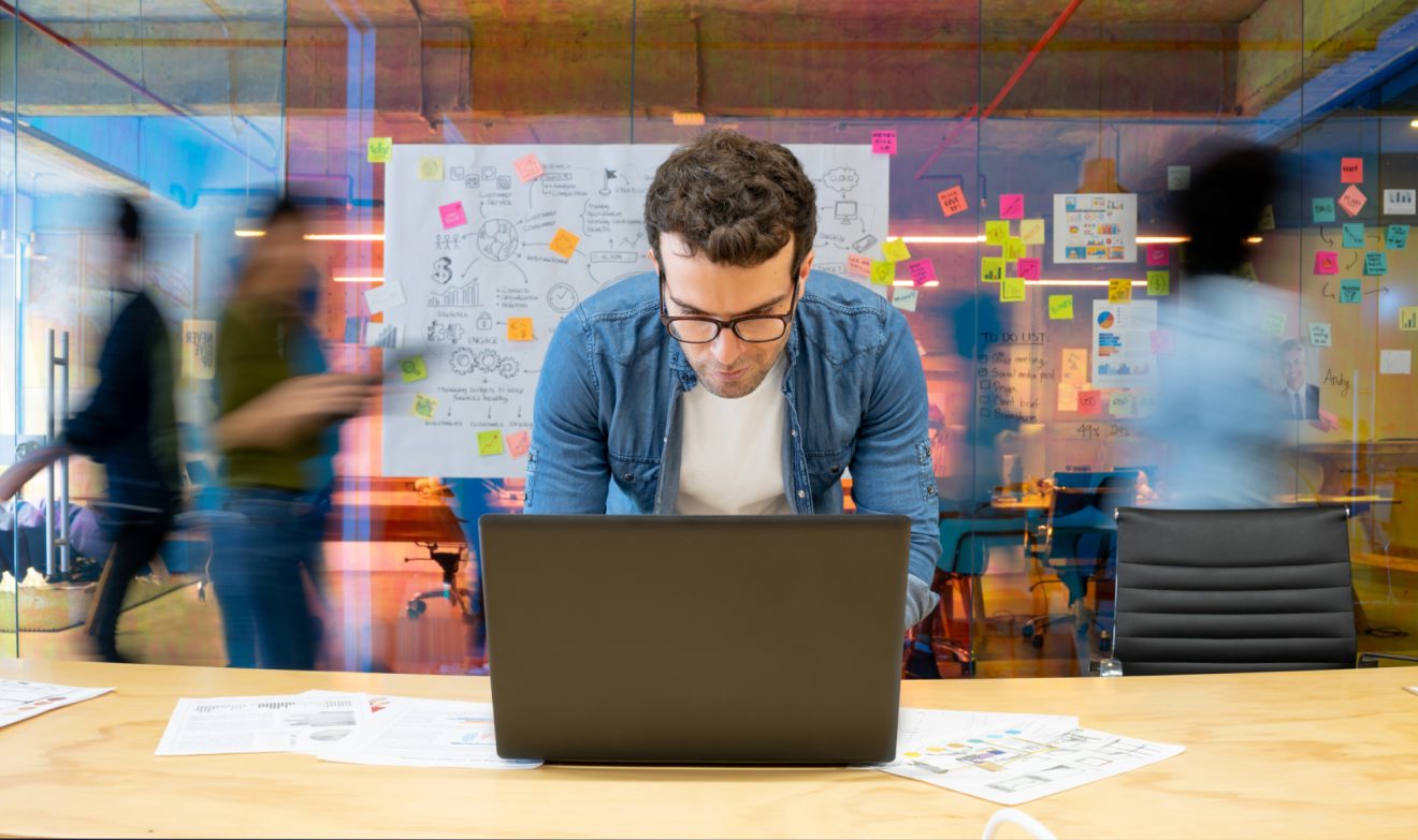 Man leaning over laptop at desk while people walk quickly behind him