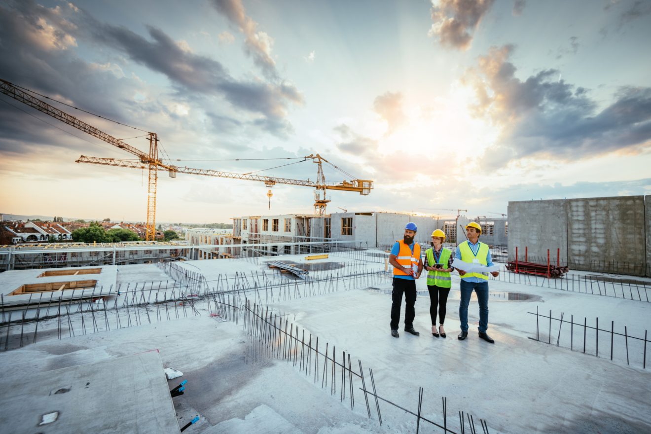 Three people in bright vests and hardhats stand on a construction site with two cranes in the background