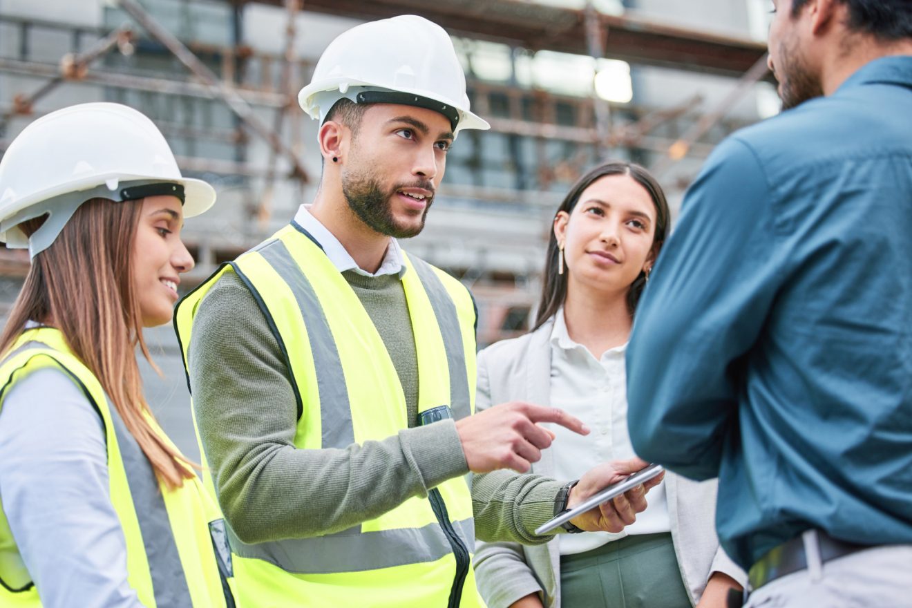 Two people in safety vests and hardhats speaking with two people in business suits