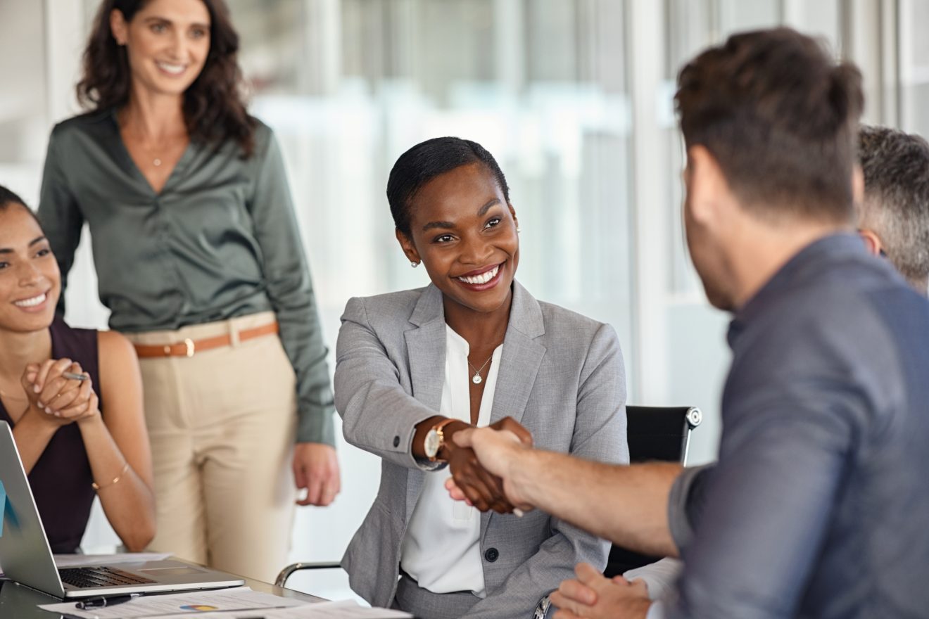 Woman and man shaking hands at a desk, surrounded by smiling onlookers