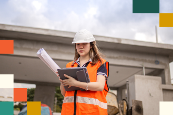 female construction worker in hard hat and reflective vest holding rolled up plans and looking at tablet computer