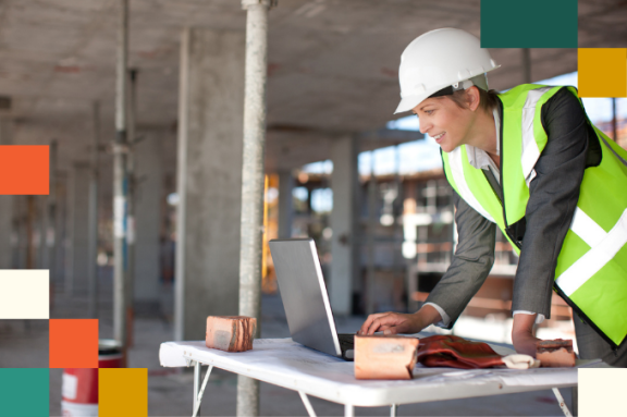 woman in hardhat and reflective vest looks at laptop on construction site